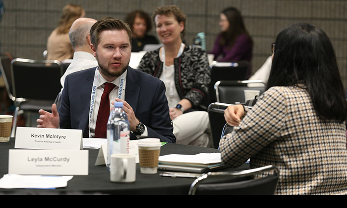 Kevin McIntyre sits at a table and chats with a woman with long dark hair and her back to the camera. 