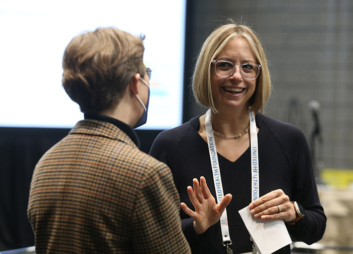 White woman with blonde bob and wearing eyeglasses smiles while talking to another person with their back to the camera. 