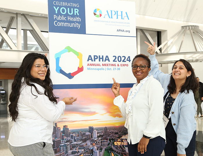 Three young adult women smile and pose in front of an APHA 2024 sign while pointing to the sign. 