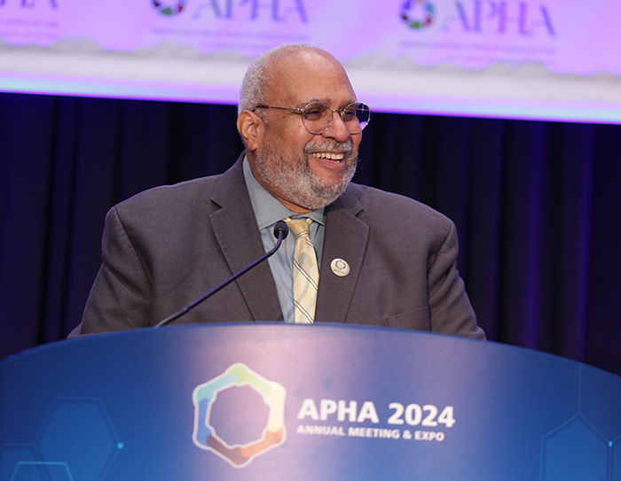 APHA Executive Director Georges Benjamin stands at a lectern with the APHA Annual Meeting on the front of it; he is smiling at the crowd. 