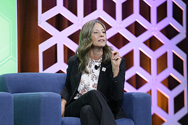 Amy Acton sits on blue chair on a stage while talking to audience. 
