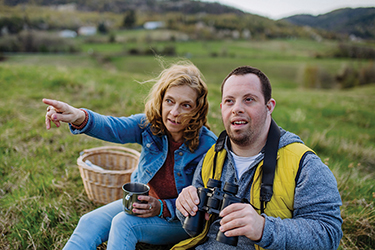 Two people sit on grass. One hold binoculars while they bird-watch. 