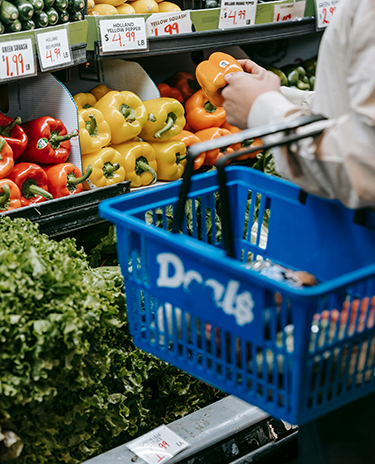 A person is holding a blue shopping basket on their left arm while selecting choosing an orange pepper in a grocery store aisle. 