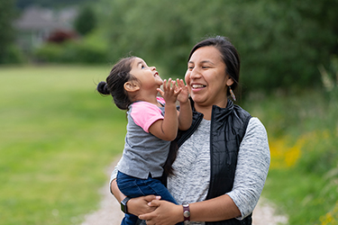 An indigenous woman holds a toddler girl in her arms outdoors with green grass and trees surrounding them. Both are laughing