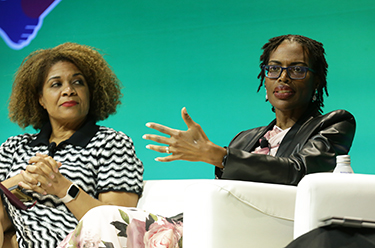 Ruqaiijah Yearby sits on a white chair on a stage talking to the audience and using her hands, while Fatima Goss Graves sits on her right. 