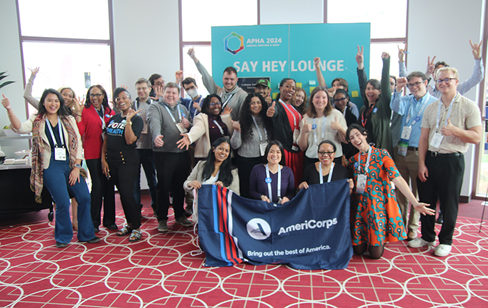 A group of about 25 pose together, smiling and laughing with some arms raised and thumbs up, while holding an Americorps banner. 