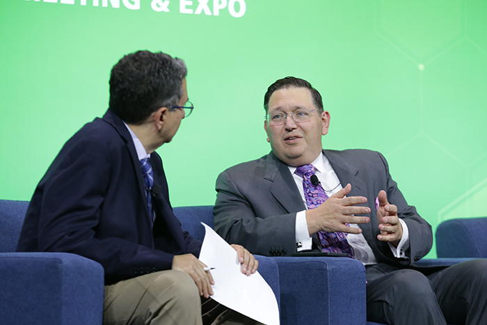 Brian Castrucci sits in a blue chair on stage talking to another man sitting next to him. 