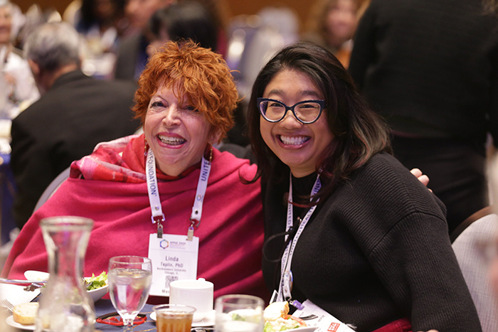 Two women sit at a round table smiling for the camera. 