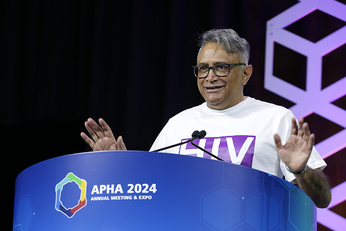 A person in a white T-shirt that says HIV Positive" in purple stands at a blue lectern and speaks to the audience. 