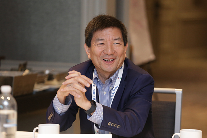 A person sits at a table with elbows on table and hands clasped together while smiling. 