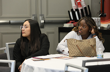 Two student members sit at a round white conference table, chairs turned while listening to a presentation. 