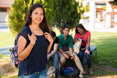 Teenage girl smiling at camera and wearing a school backpack, with other students sitting on bench behind her. 