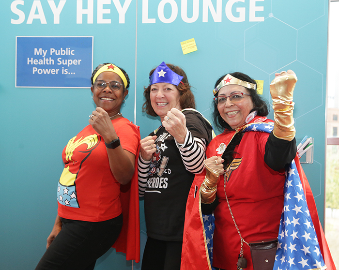 Three people dressed as women superheroes smile and pose with their fists toward the camera. 