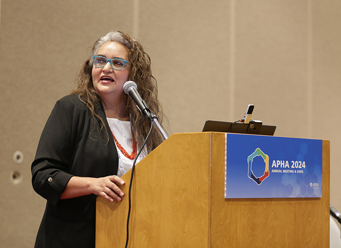 A woman stands at a lectern in front of a microphone and looks off camera toward her slides while talking. 