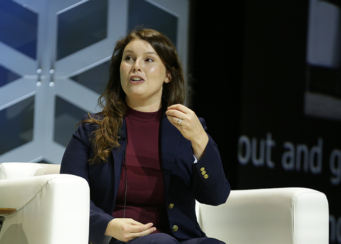 A person sits on a white cozy chair on a stage and talks to the audience using hand gestures. 