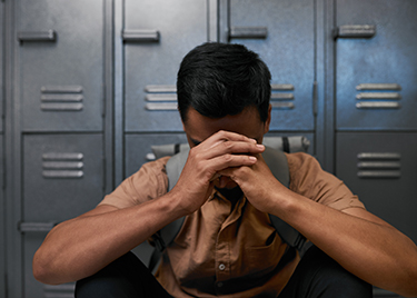 Man sits aginst a set of gray lockers, with his elbows resting on his knees and hands clasped together and head resting on hands, with his face hidden.