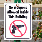 A white sign that states No Weapons Allowed Inside This Building with a photo of a handgun with a crossed-out circle around it. 