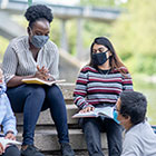 A group of teenagers sits on steps outdoors with open notebooks as they talk. 