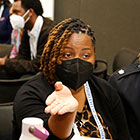 A woman with braids and wearing a black masks moves her hands to make a point. 